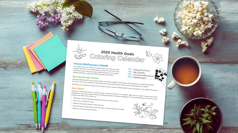 2025 Health Goal Coloring Calendar on a desk surrounded by pens, sticky notes, and healthy food. 