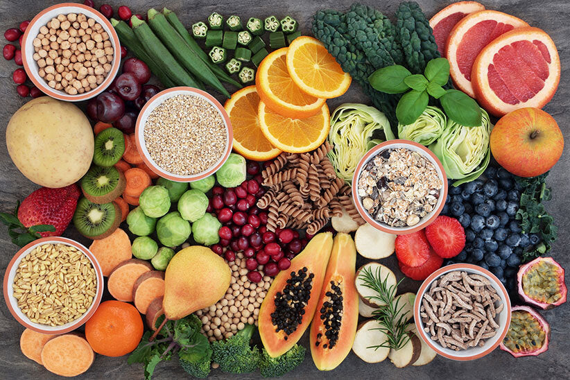 Top down photo of high fiber foods on a counter top, including whole wheat pasta, fruits, vegetables, seeds, nuts, and more